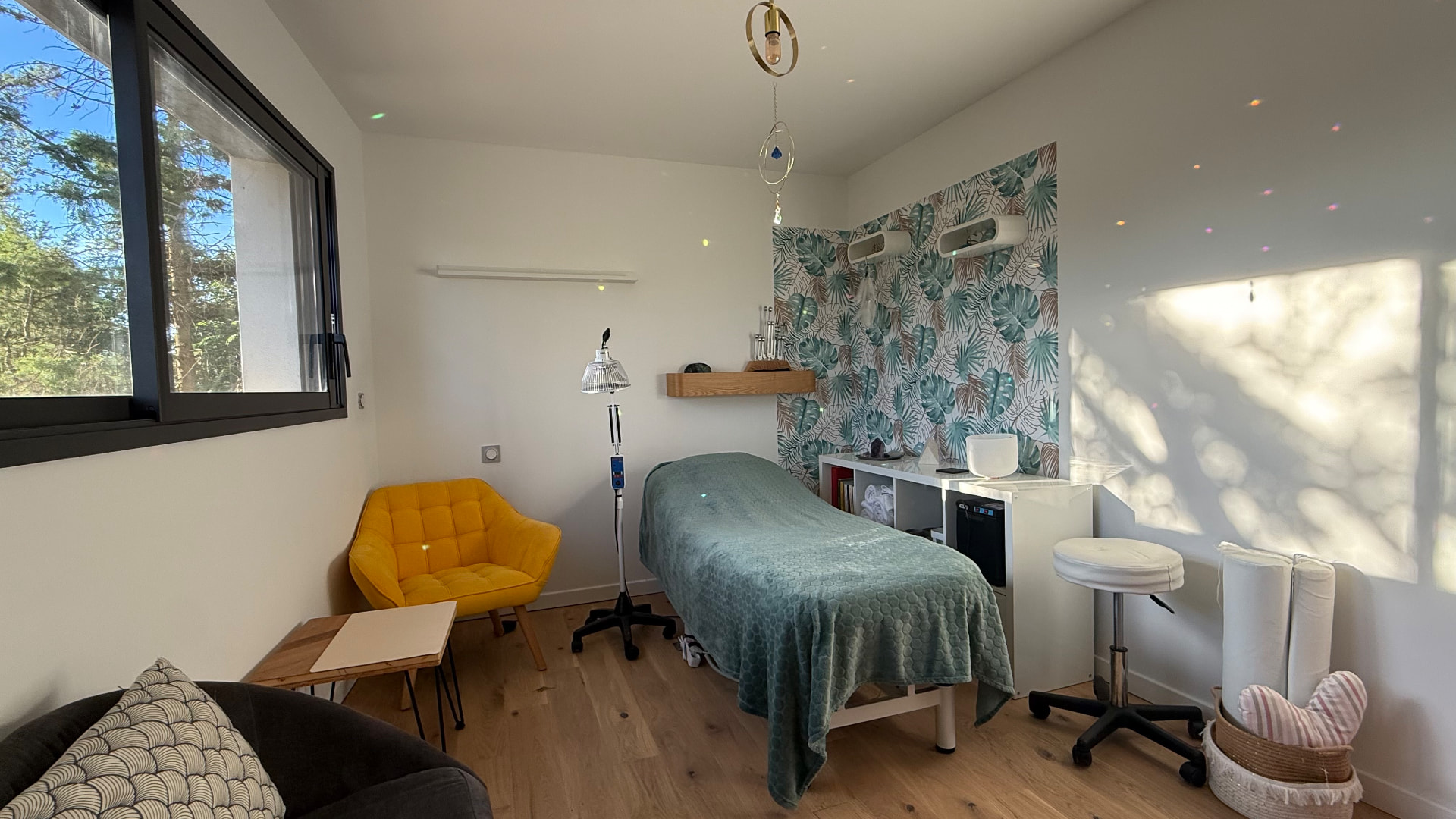 La salle de soin lumineuse avec table de réflexologie, fauteuil jaune et grande fenêtre sur les arbres